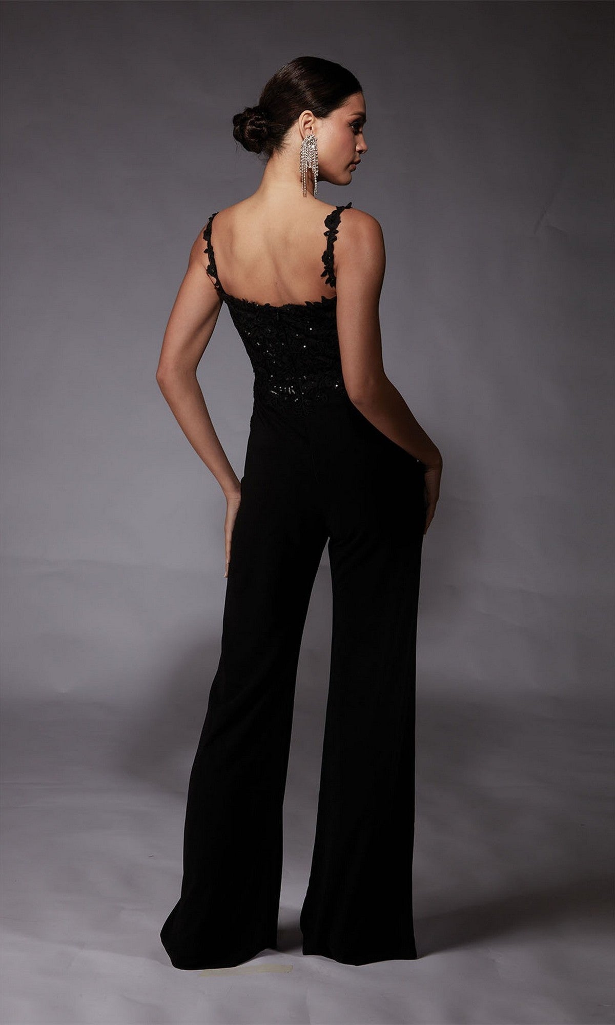 A woman stands with her back to the camera, wearing the Alyce 70073 Long Formal Jumpsuit featuring lace straps and wide-leg pants, paired with large dangling earrings.
